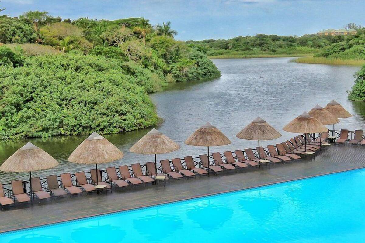 Une piscine bordée de chaises longues et ombragée par des parasols en chaume, le tout entouré d’une végétation luxuriante et d’une rivière.