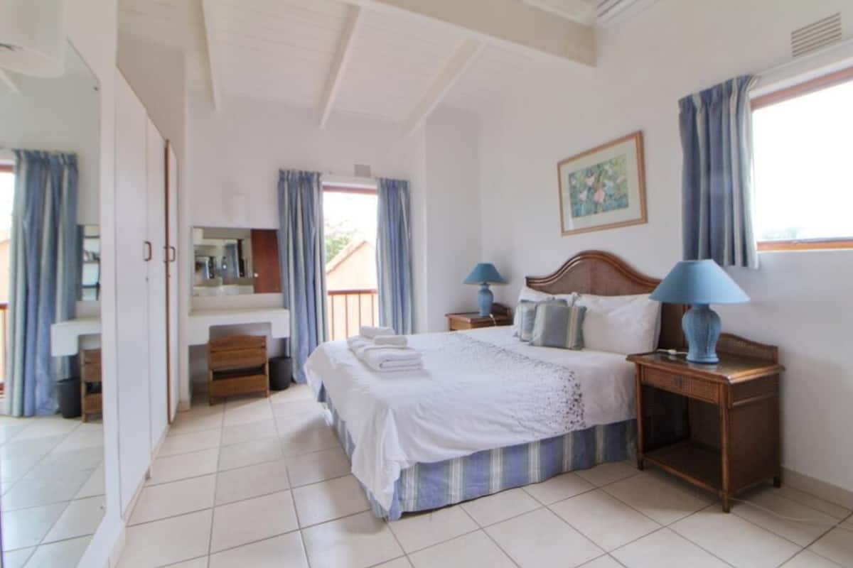 Un dormitorio con cama, dos mesitas de noche con lámparas, un aparador, un espejo y una ventana con cortinas.