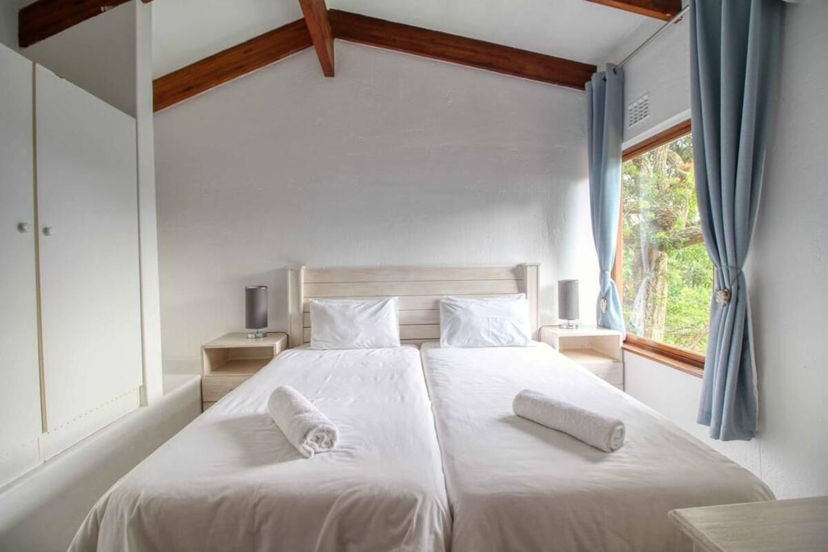 Un lit double avec du linge de lit blanc, une tête de lit en bois, des tables de chevet avec des lampes et une fenêtre avec des rideaux.