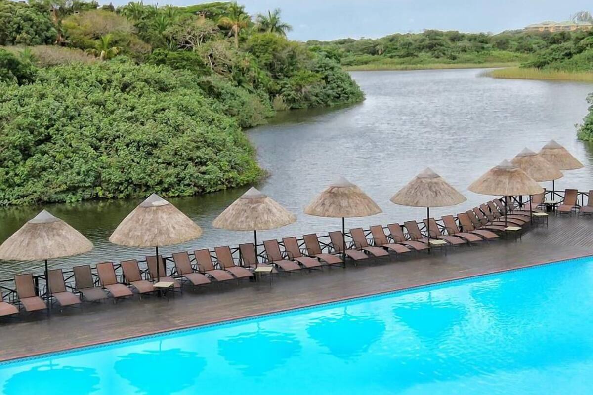 Une piscine bordée de chaises longues et ombragée par des parasols en chaume, le tout entouré d’une végétation luxuriante et d’une rivière.