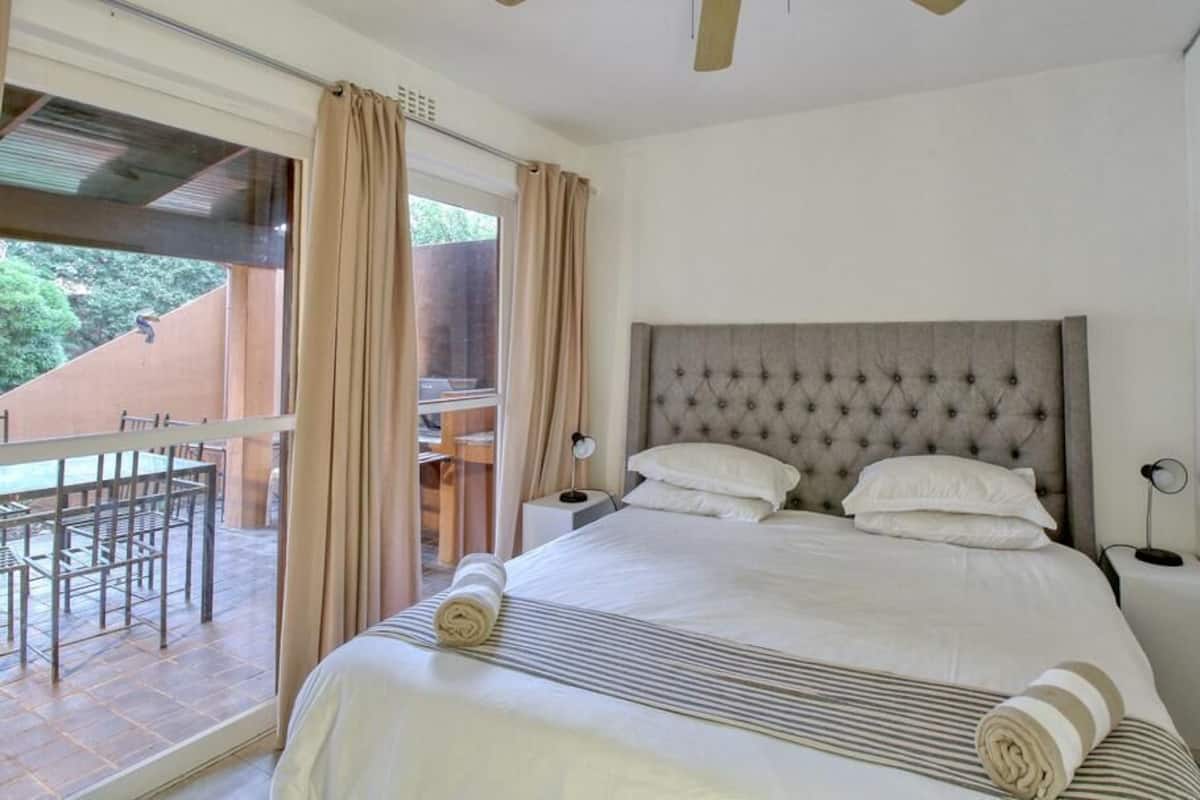 Une chambre avec un grand lit, une table de chevet, une commode, une fenêtre avec des rideaux et une vue sur une terrasse extérieure.