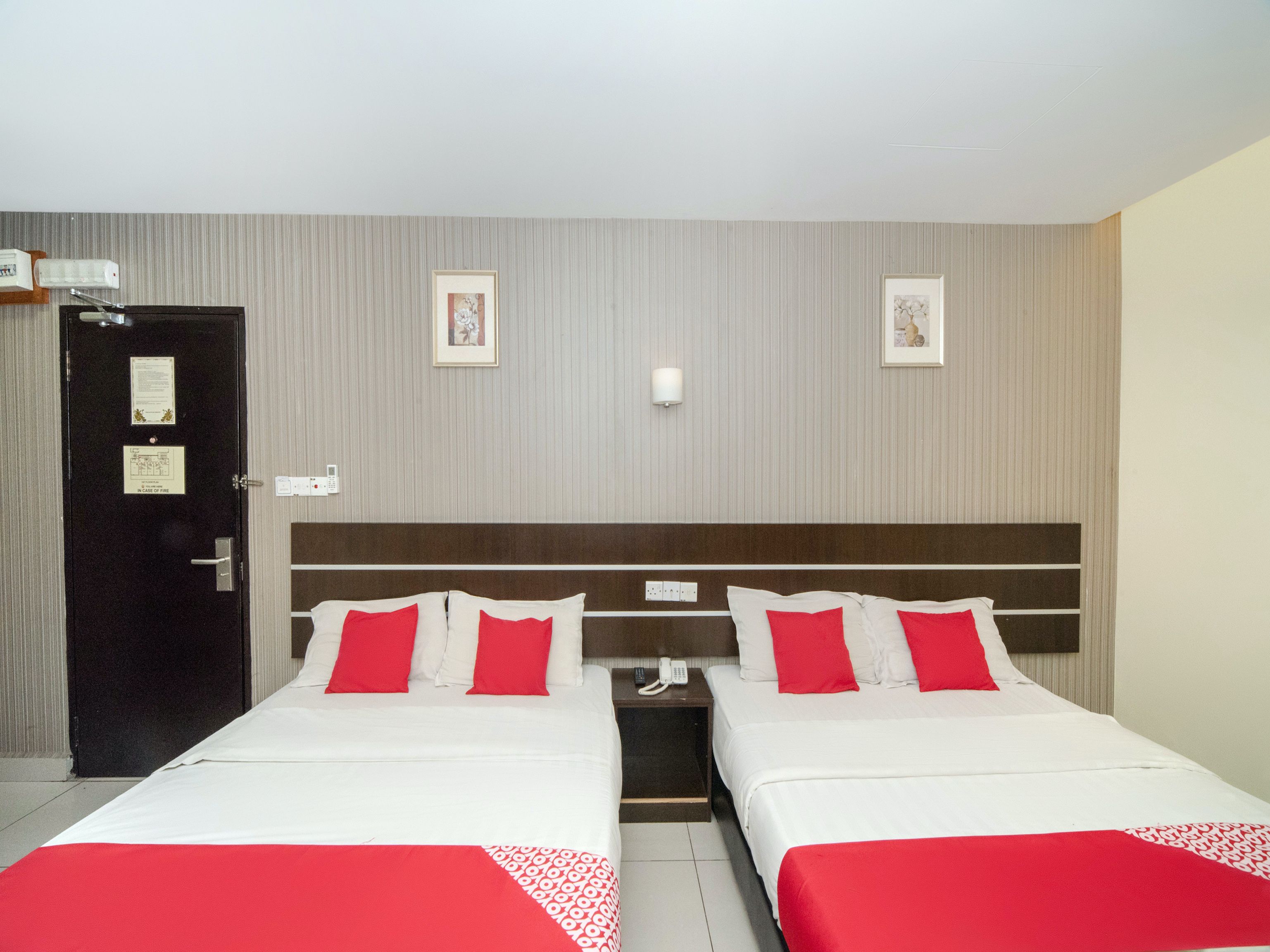 A hotel room with two beds, a door with a fire safety sign, a telephone, and two framed pictures on the wall.