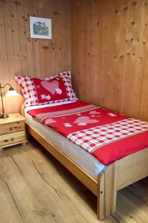 Ein Einzelbett mit einer rot-weißen Karodecke, einem Holz-Kopfstück und einem Nachttisch mit Lampe.