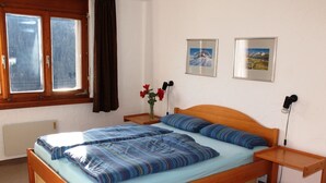 Un dormitorio con una cama de madera, una mesita de noche, un escritorio junto a la cama, una ventana con cortinas y dos cuadros en la pared.