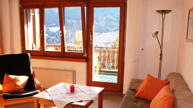Una habitación con una silla de madera, una mesa con mantel blanco, un sofá con cojines naranjas y vistas a montañas a través de grandes ventanales.