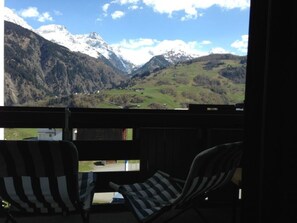 Ein Balkon mit zwei gestreiften Stühlen, der auf eine gebirgige Landschaft mit schneebedeckten Gipfeln und klarem Himmel blickt.