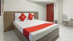 A modern hotel room with a bed, two red pillows, a red bedspread, a wooden headboard, a white wall, a red door, a white chair, and a small table.