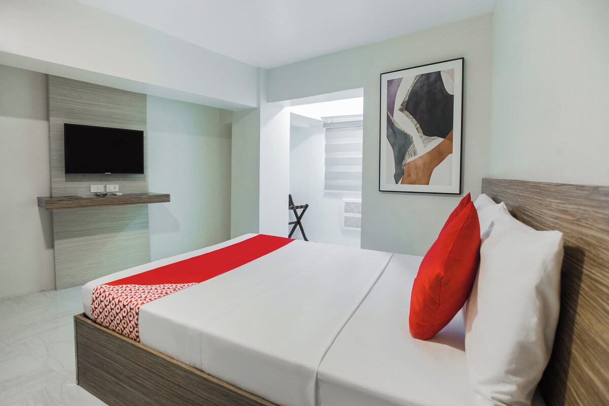 A modern hotel room with a large bed, a flat-screen TV mounted on the wall, a wooden bedside table, and a framed abstract artwork.