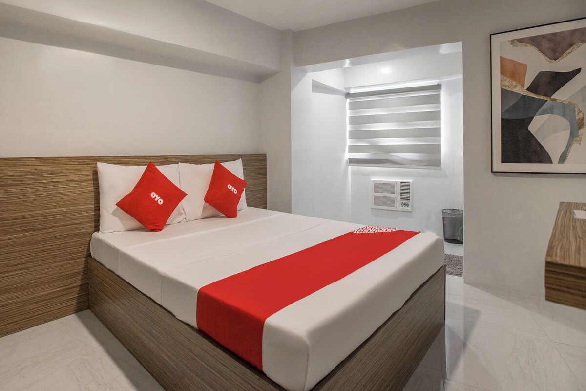 A modern hotel room with a large bed, wooden headboard, and a red accent on the bedspread.
