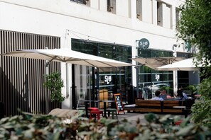 An outdoor seating area with tables and chairs, a signboard, and a building with large windows. - Slo Hostel (Bordeaux)