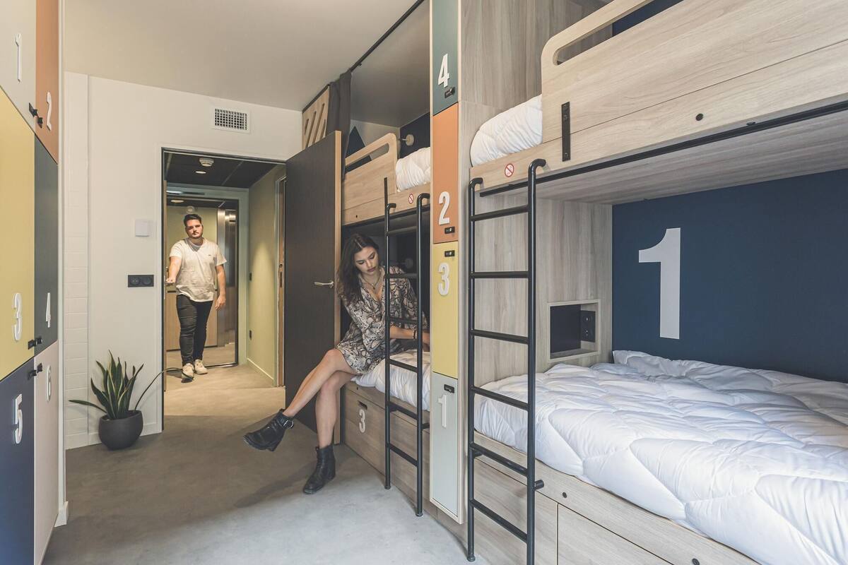 A dormitory-style room with bunk beds, a door with a number, and a potted plant.