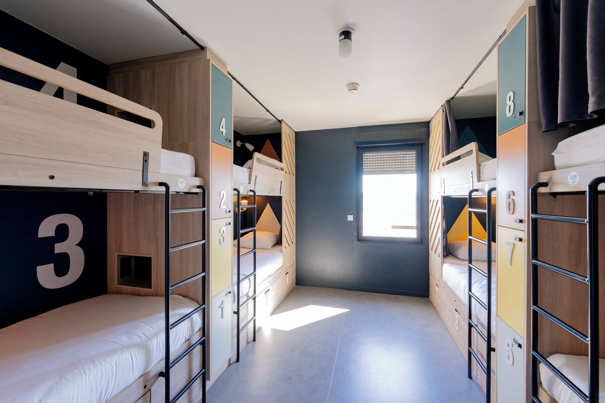A dormitory-style room with bunk beds, numbered lockers, and a window providing natural light.