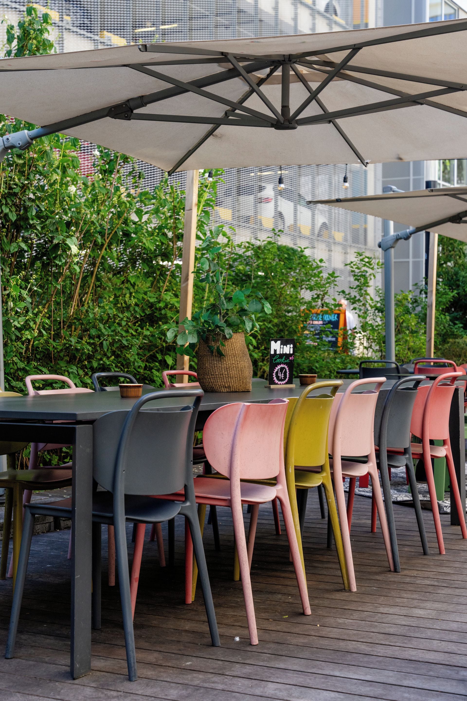 An outdoor seating area with tables and chairs, a signboard, and greenery in the background.