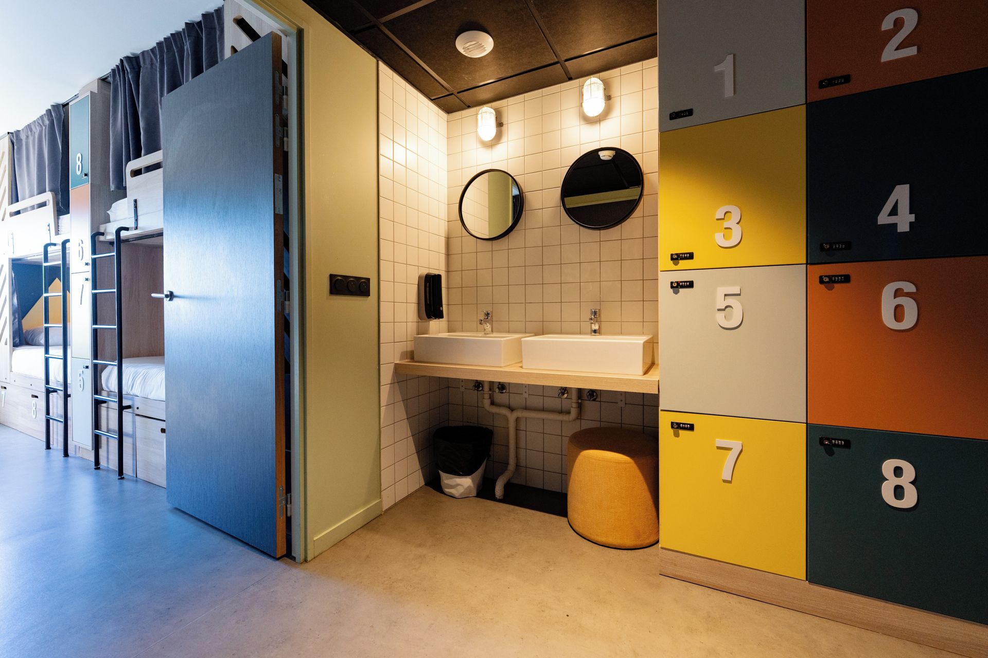 A hotel bathroom with bunk beds, a sink, two mirrors, and lockers numbered 1 to 8.