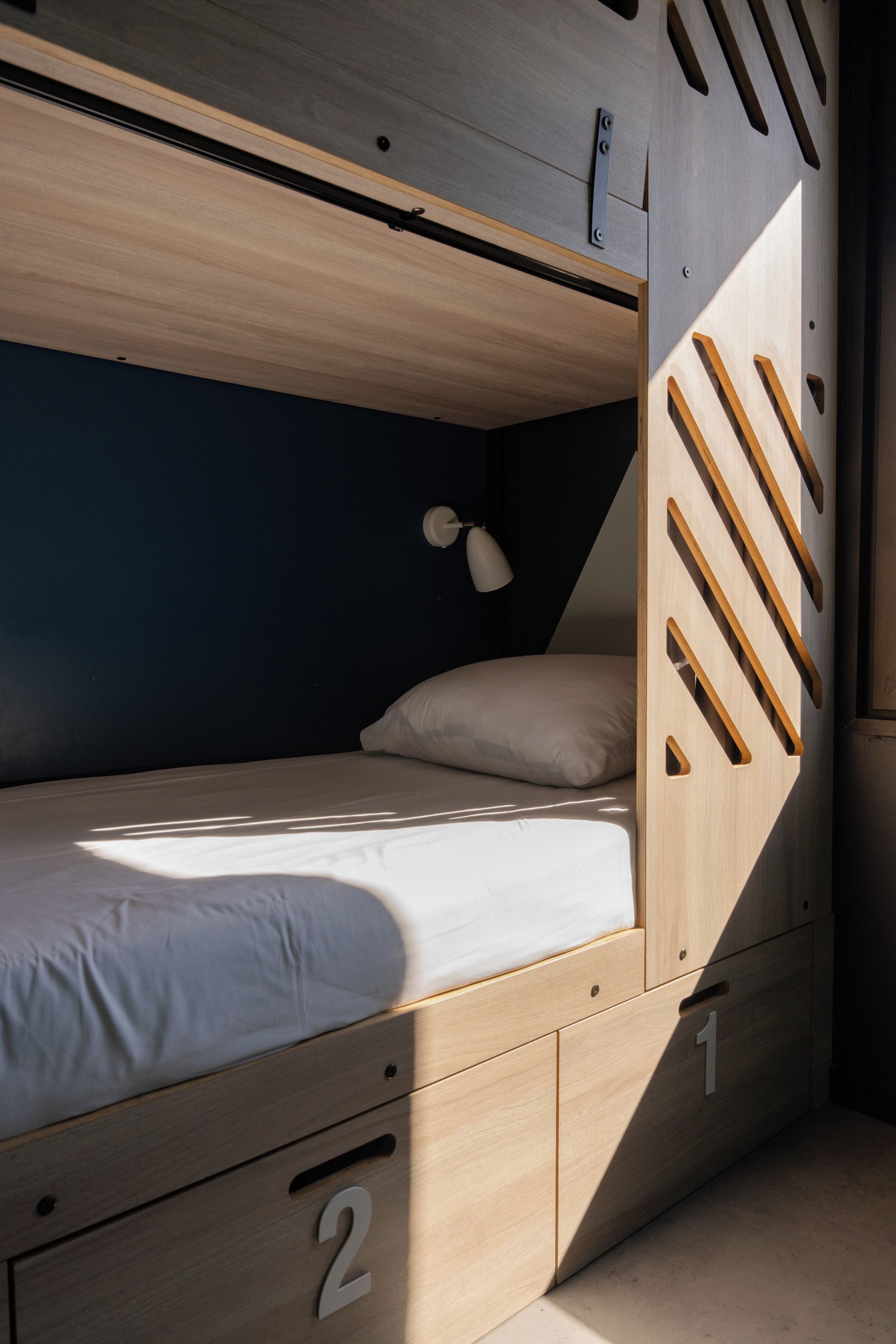 A bunk bed with a wooden ladder, a single bed with a white pillow, and a small desk with drawers numbered 1 and 2.