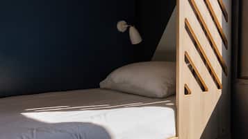 Una cama litera con escalera de madera, una cama individual con almohada blanca y un escritorio pequeño con cajones numerados 1 y 2.