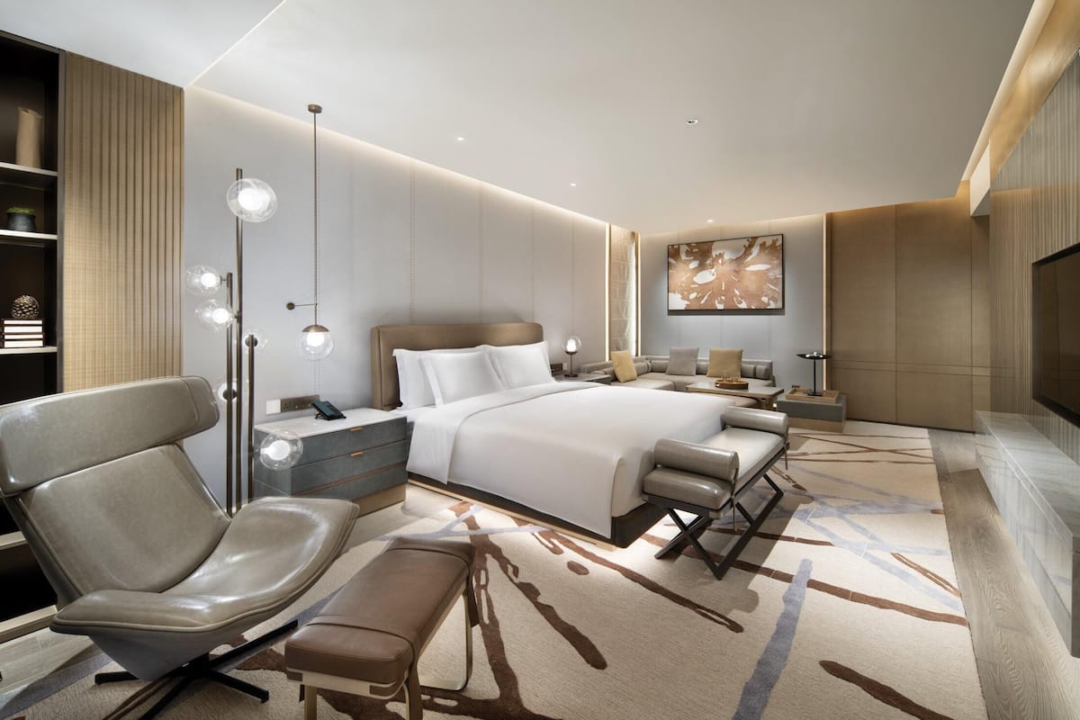 Una habitación de hotel moderna con una cama grande, una zona de estar con una silla y un banco, una mesita de noche, una lámpara de pie y una obra de arte de gran tamaño en la pared.