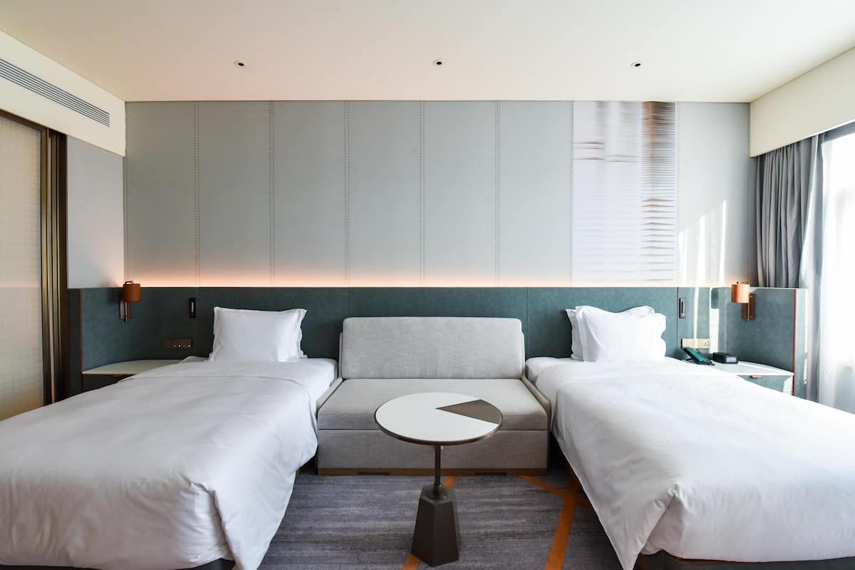 Habitación de hotel con dos camas, un sofá, una mesita redonda y un ventanal con cortinas.