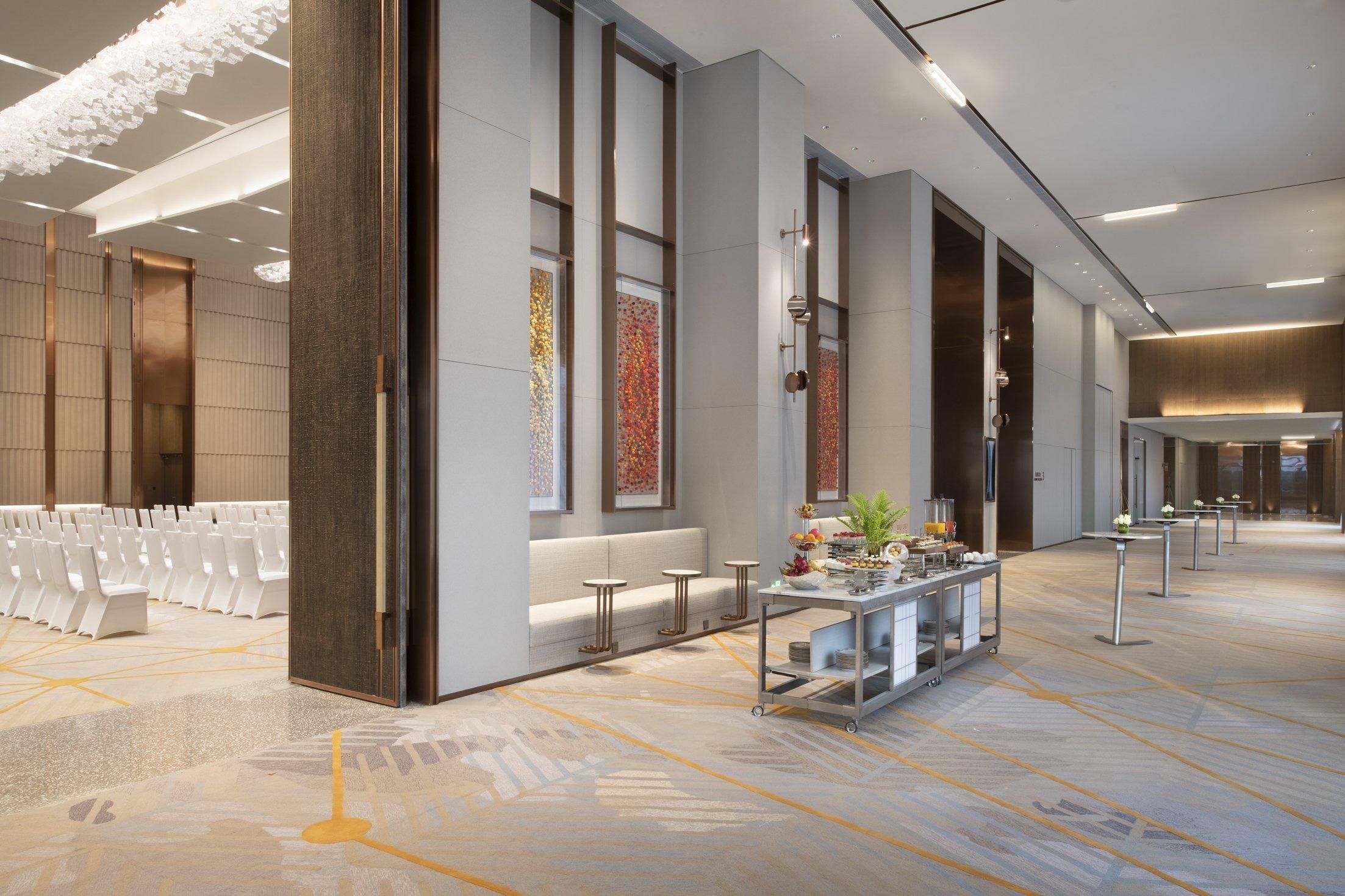 A spacious hotel lobby with rows of white chairs, a reception desk, and a decorative wall with patterned artwork.