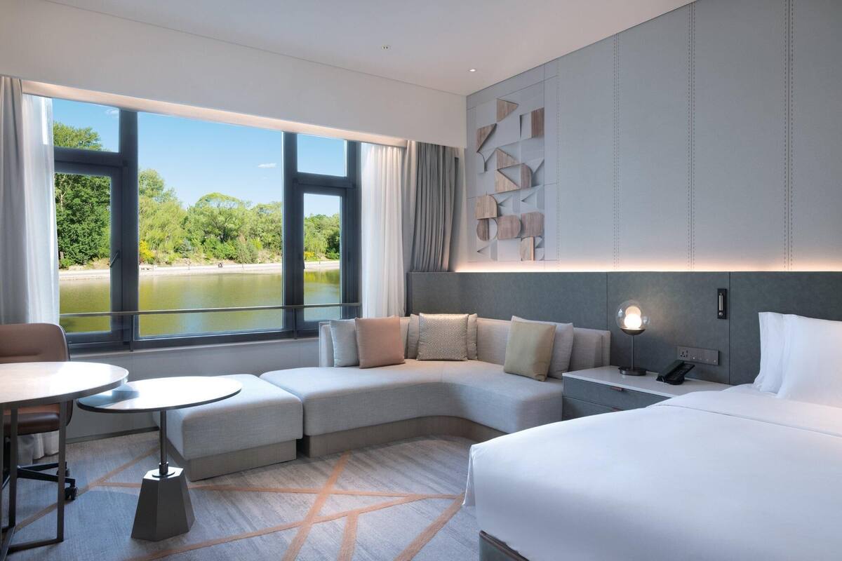 Una habitación de hotel moderna con un sofá curvo, una pequeña mesa redonda, una cama y vistas a un lago.