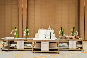 A banquet table with a tiered cake and white orchids, set against a plain wall with a tiled floor.