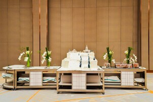 A banquet table with a tiered cake and white orchids, set against a plain wall with a tiled floor.