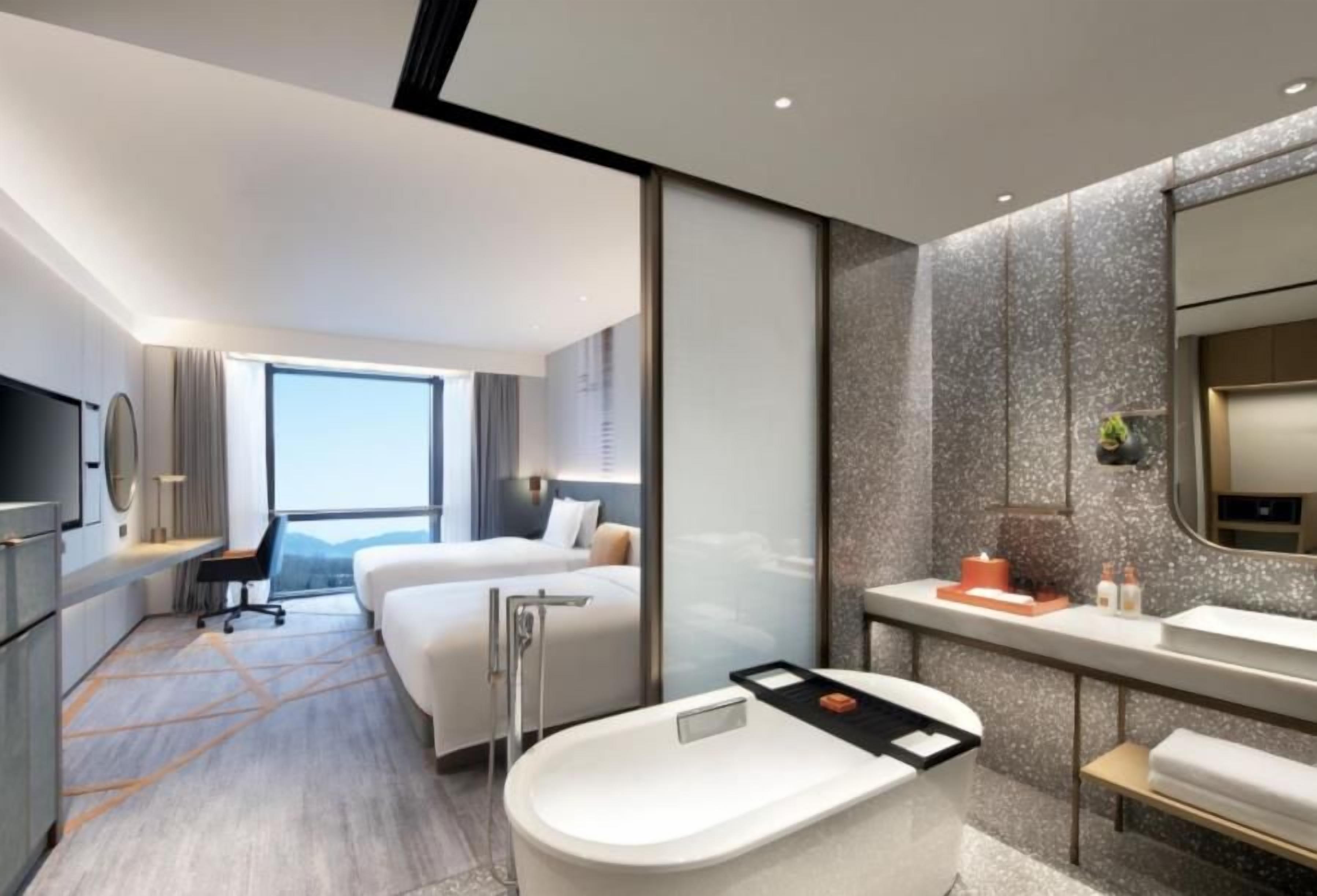 A modern hotel room with a large bed, a separate desk area, a bathroom with a bathtub, and a large window providing a view.