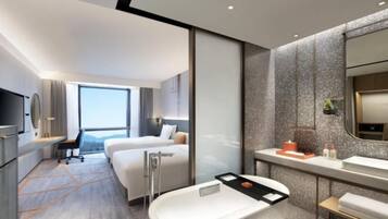 A modern hotel room with a large bed, a separate desk area, a bathroom with a bathtub, and a large window providing a view.