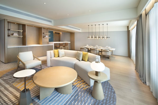 Una habitación de hotel moderna con sofá, dos mesas redondas, zona de comedor y estanterías empotradas.