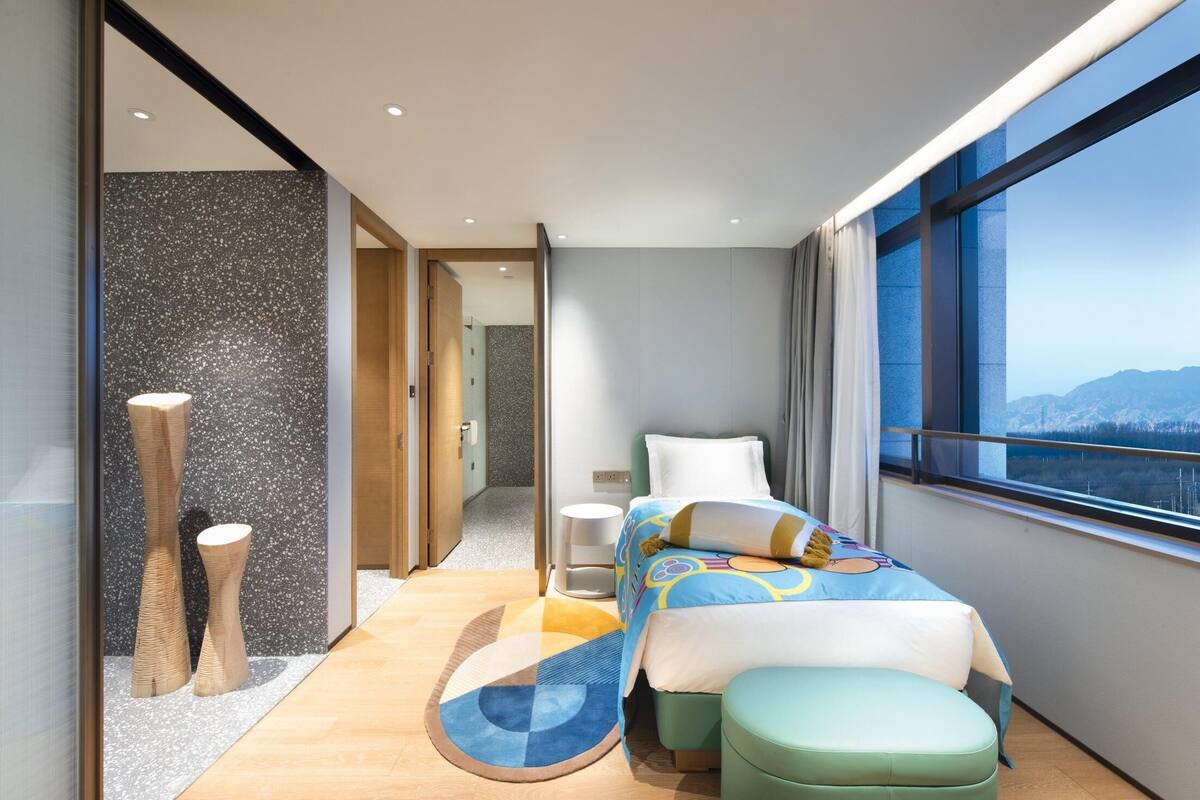Habitación de hotel moderna con una cama, un taburete pequeño, un tapete redondo y vistas a las montañas a través de amplias ventanas.