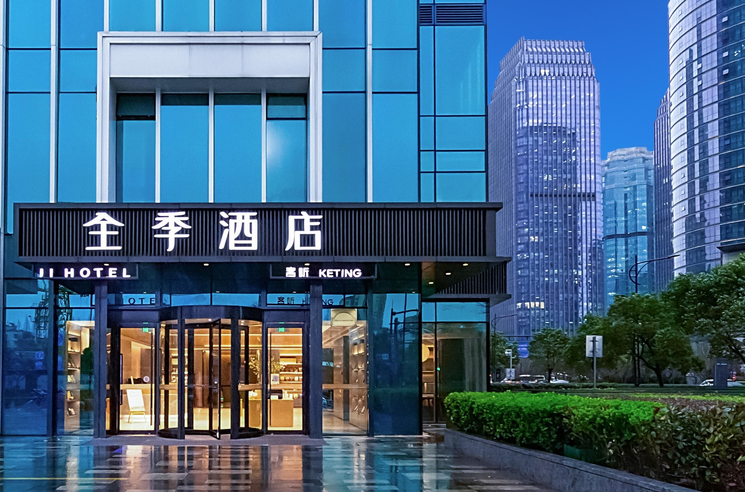 A hotel entrance with glass doors, signage in two languages, and modern urban buildings in the background.
