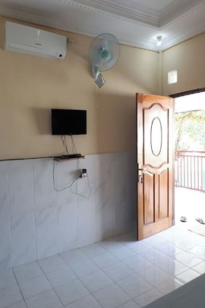A room with a tiled floor, a mounted television, an air conditioning unit, a fan, and a wooden door. - Sagita Bungalow by Zuzu (Gili Trawangan)