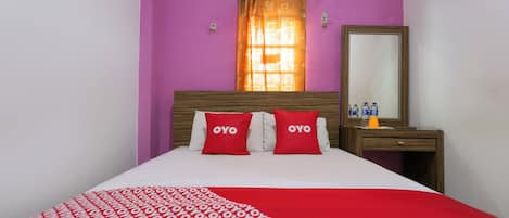 A bed with a red and white patterned bedspread, two red pillows with the word "OYO" on them, a wooden headboard, a mirror on a wooden nightstand, and a window with orange curtains.