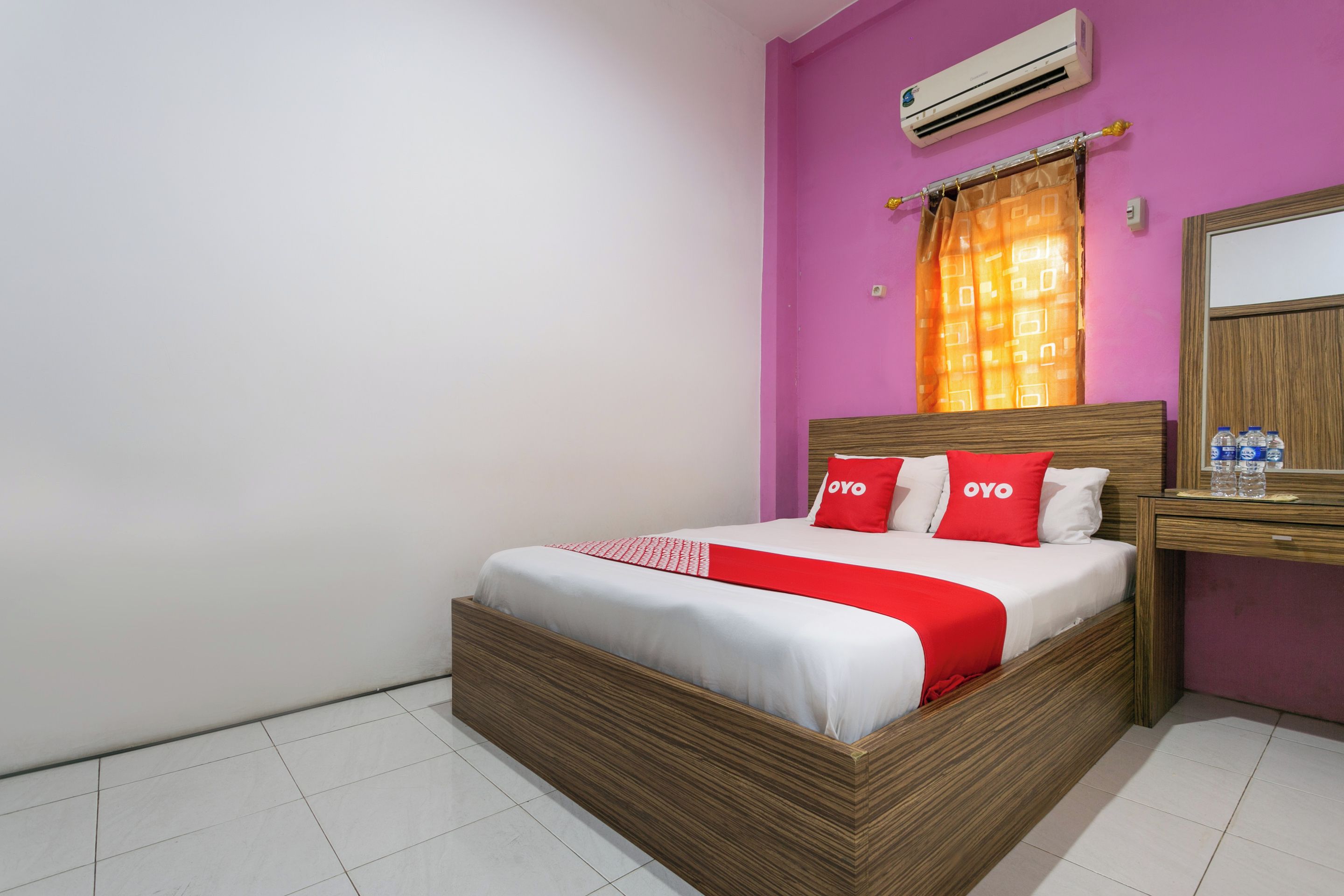A hotel room with a wooden bed, a nightstand with bottled water, a mirror, and an air conditioner on the wall.