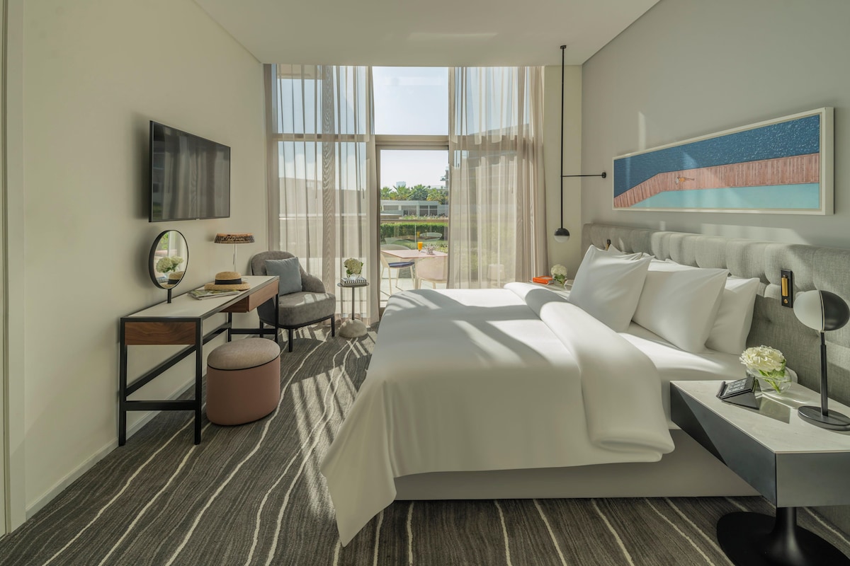 A modern hotel room with a large bed, a desk with a chair, a small table with a lamp, and a large window with curtains.