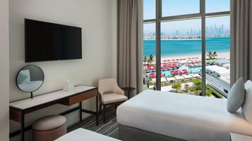 A hotel room with a bed, desk, chair, TV, and a large window with a city and beach view.