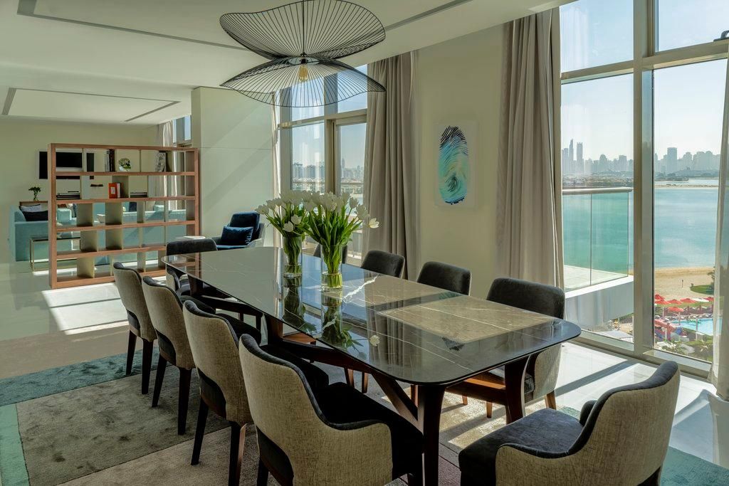 A modern dining area with a glass table, chairs, a bookshelf, and a view of the city and water.