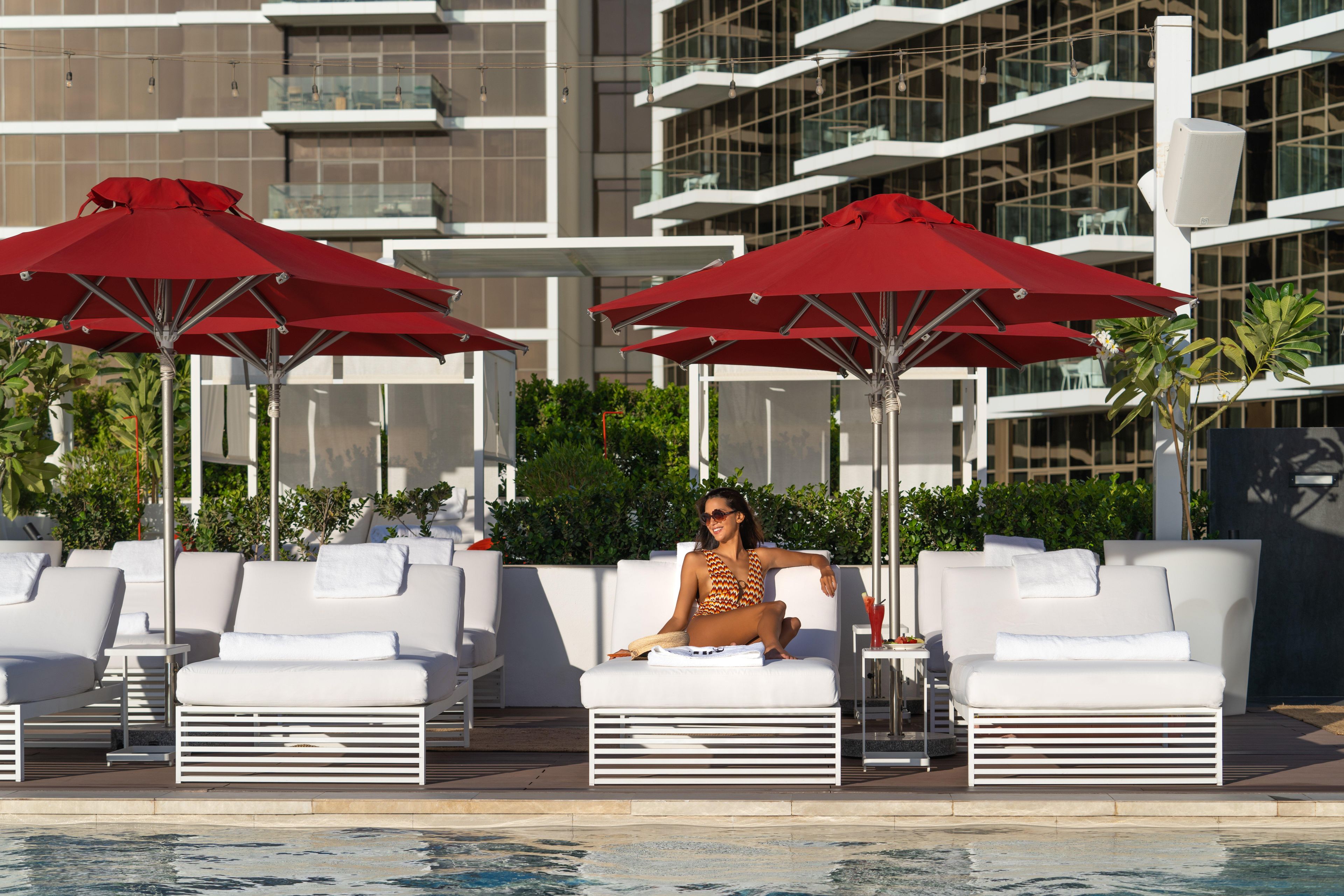 A poolside area with lounge chairs and red umbrellas, a pool, and a modern building in the background.