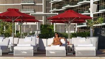 A poolside area with lounge chairs and red umbrellas, a pool, and a modern building in the background.