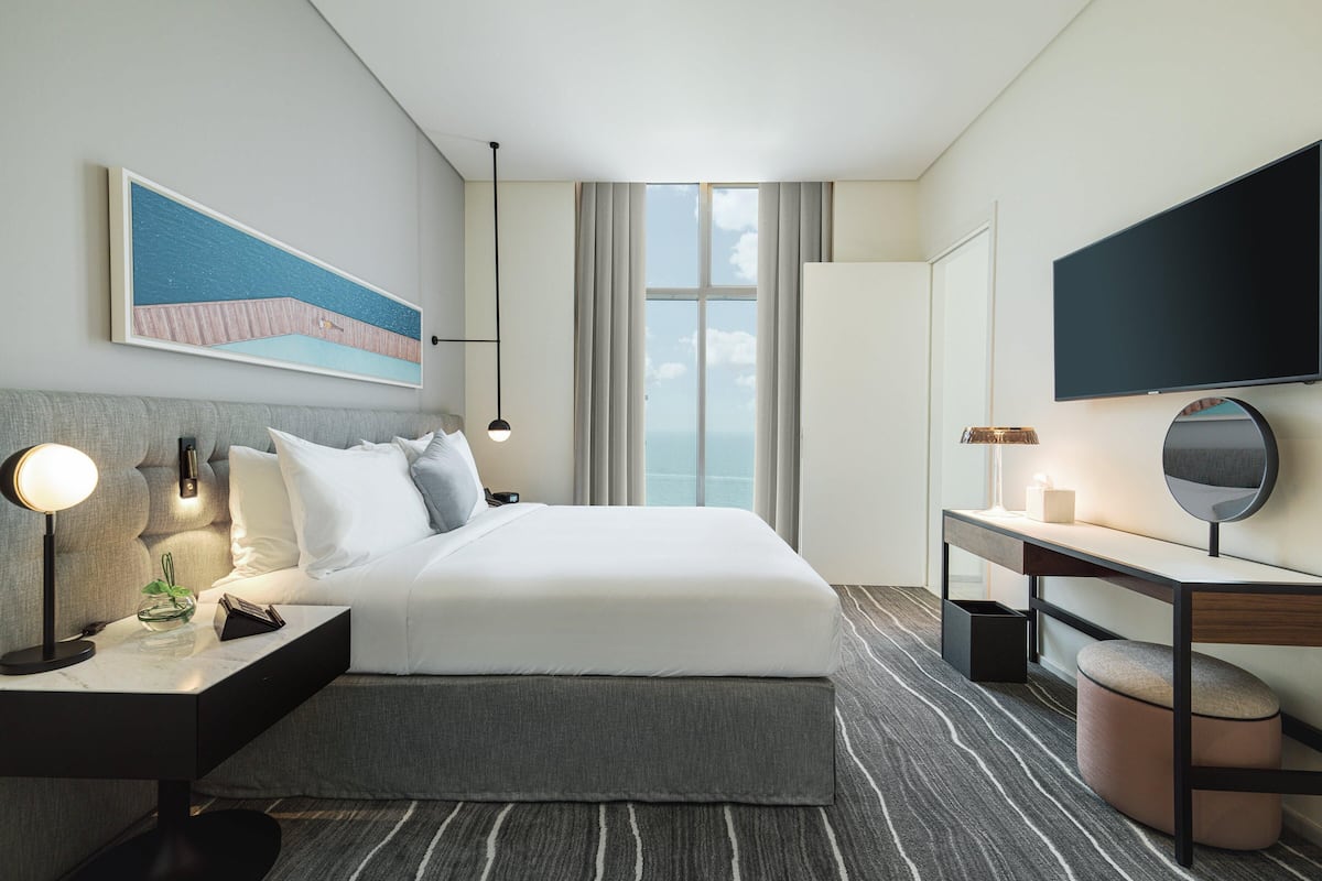A modern hotel room with a large bed, bedside tables, a desk, a flat-screen TV, and a large window offering a view.