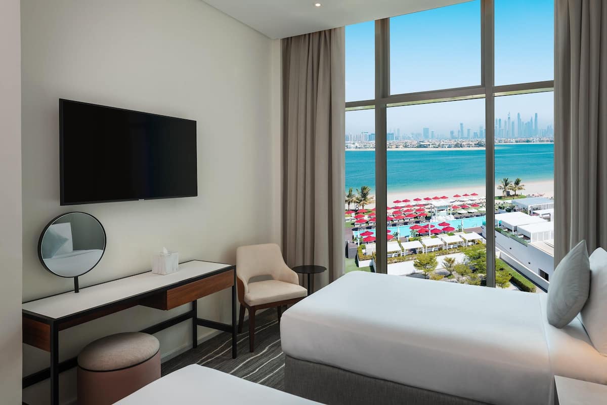A hotel room with a bed, desk, chair, TV, and a large window with a city and beach view.