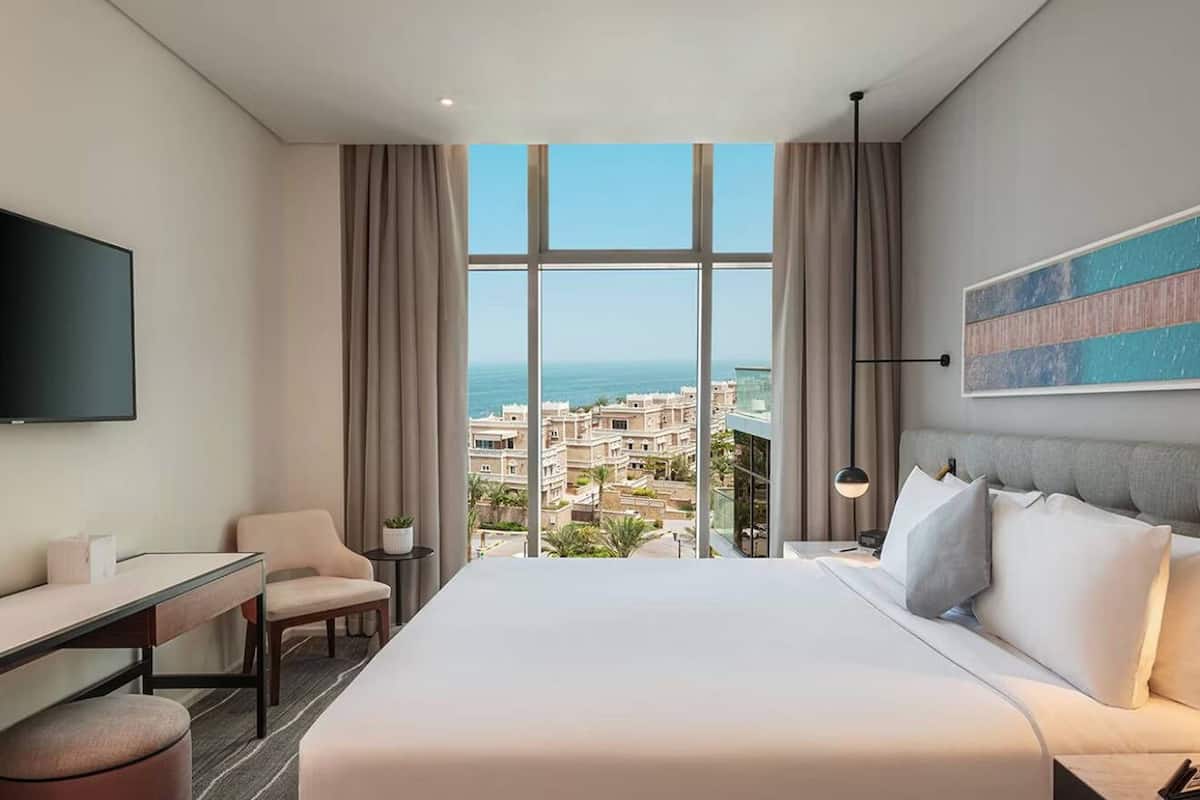 A hotel room with a large bed, a desk, a chair, a TV, and a view of the city and sea.