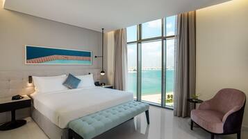 A modern hotel room with a large bed, a bench, a chair, a nightstand, a lamp, a painting, and a view of the sea.
