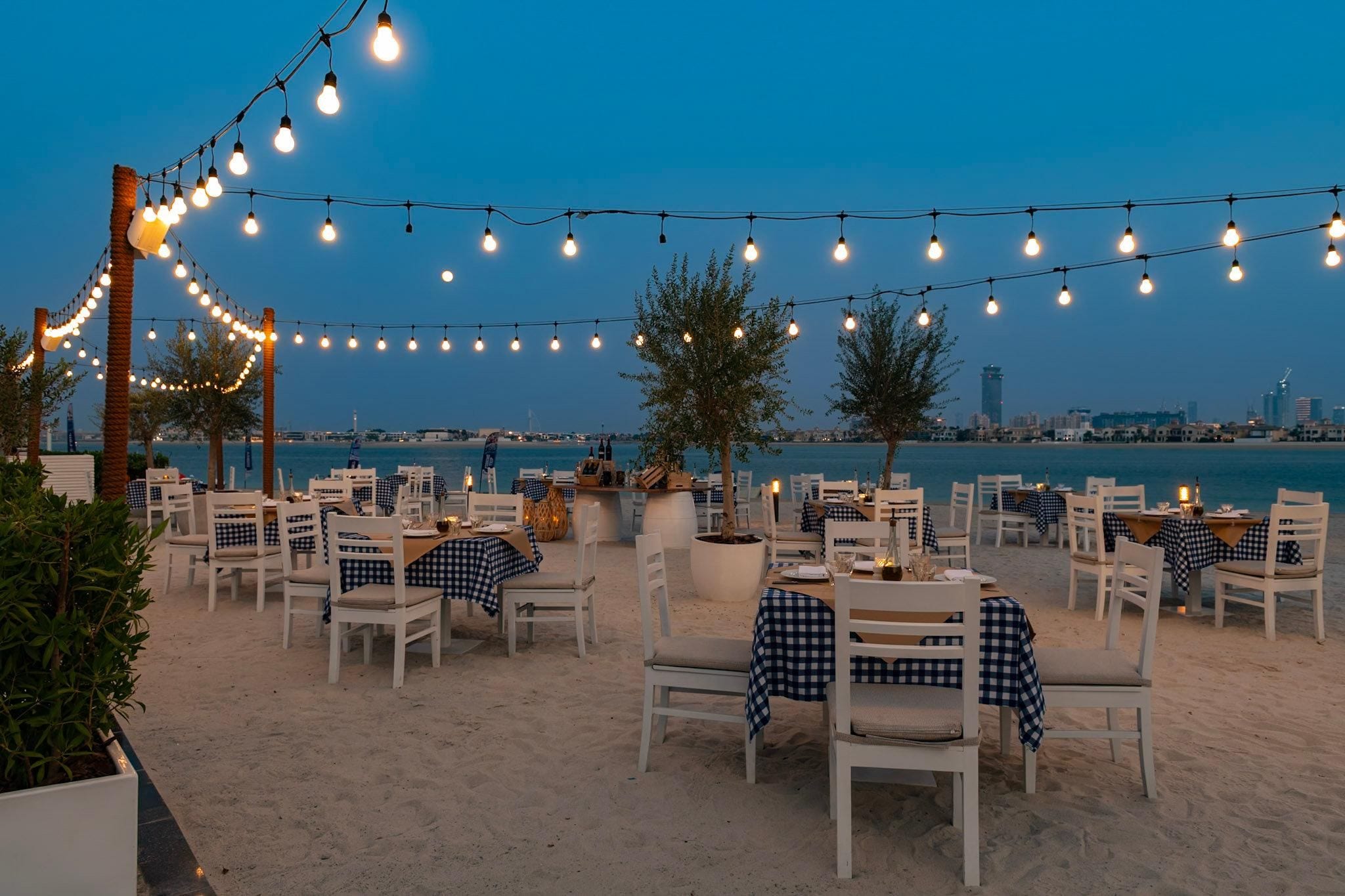 An outdoor dining area with tables and chairs, string lights, and a view of the city skyline.