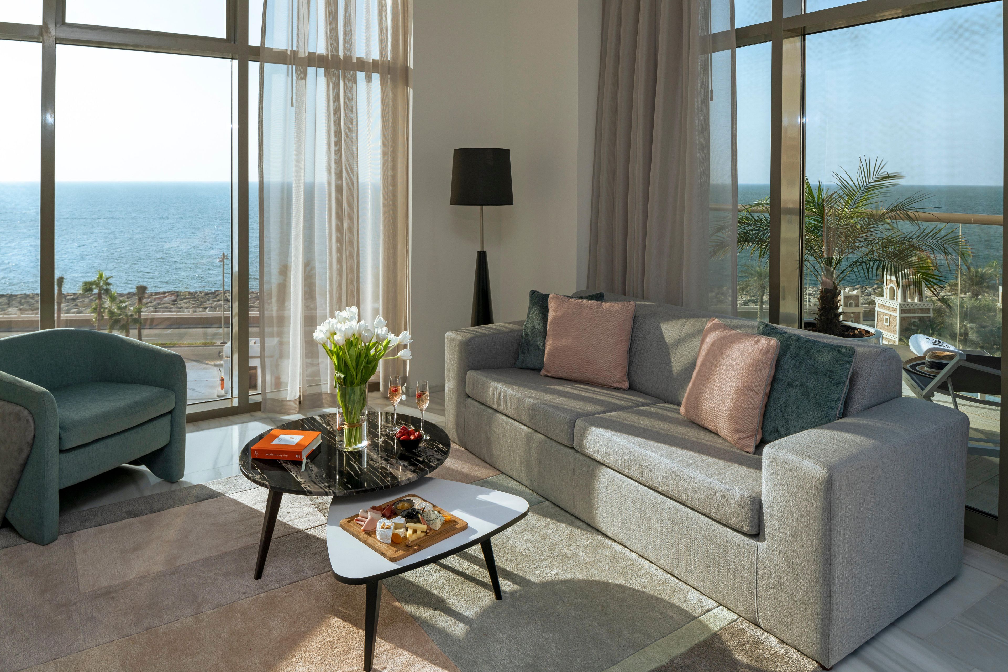 A modern living room with a sofa, a coffee table with a fruit platter, a vase with flowers, and a view of the sea.