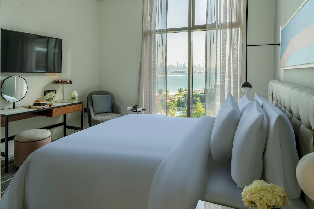 A hotel room with a large bed, a desk with a chair, a TV, and a view of a cityscape through the window.