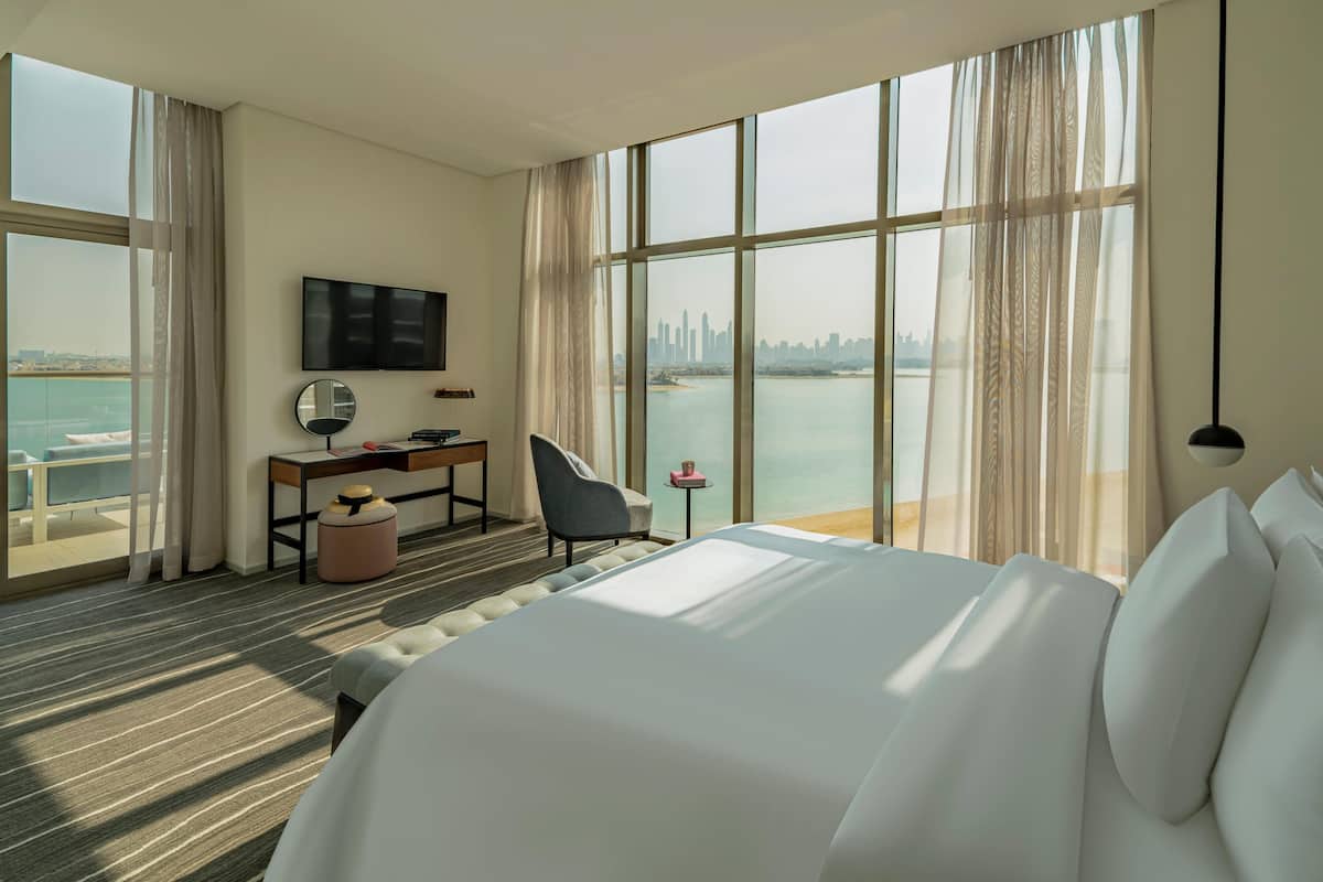 A hotel room with a large bed, a desk, a chair, a TV, and a view of the city skyline and water.