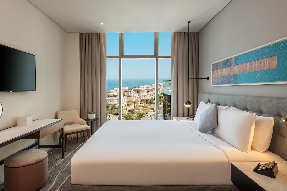 A hotel room with a large bed, a desk, a chair, a TV, and a view of the city.