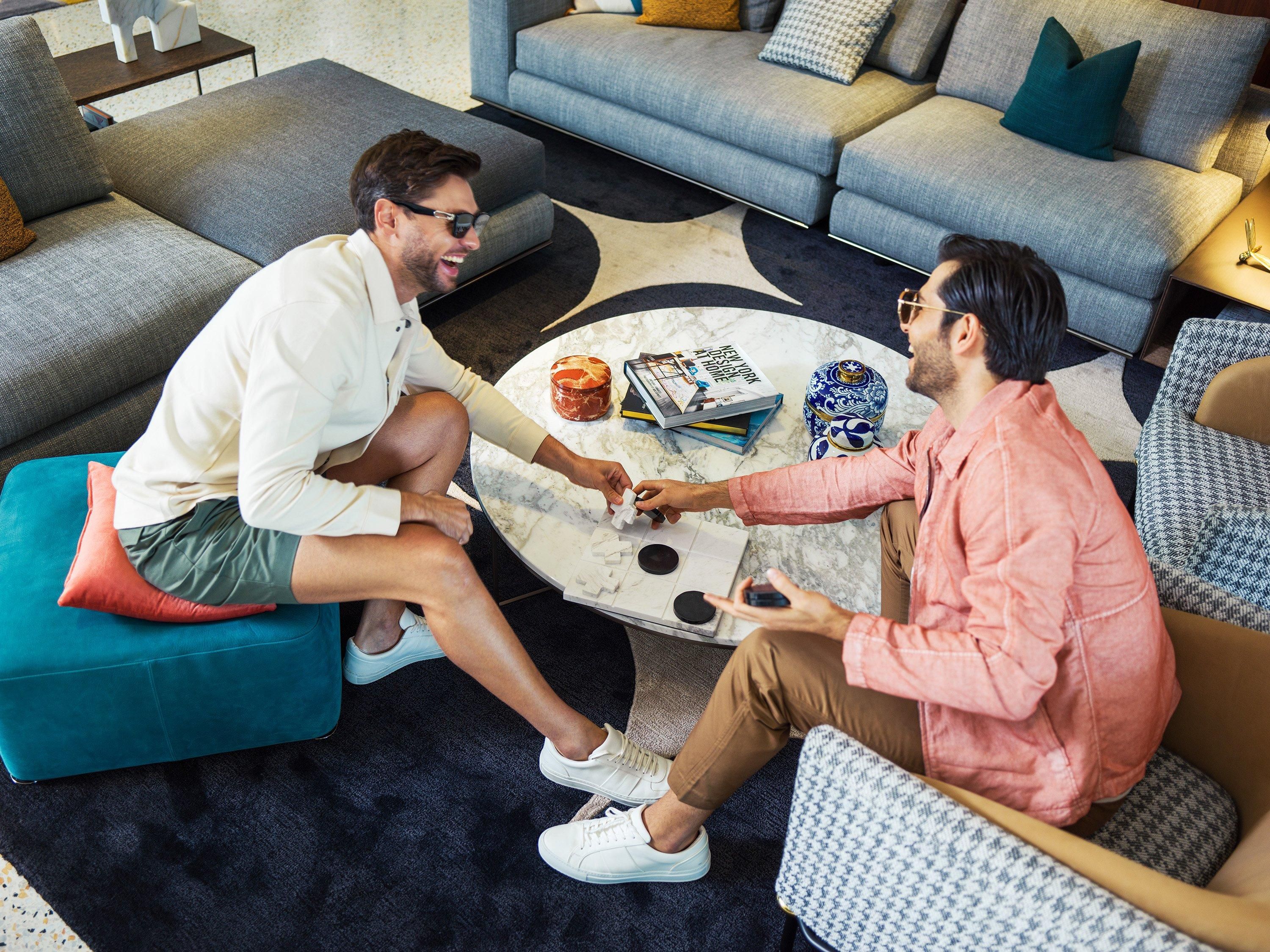 Two people seated on a round coffee table in a modern living room with a sofa and patterned rug.