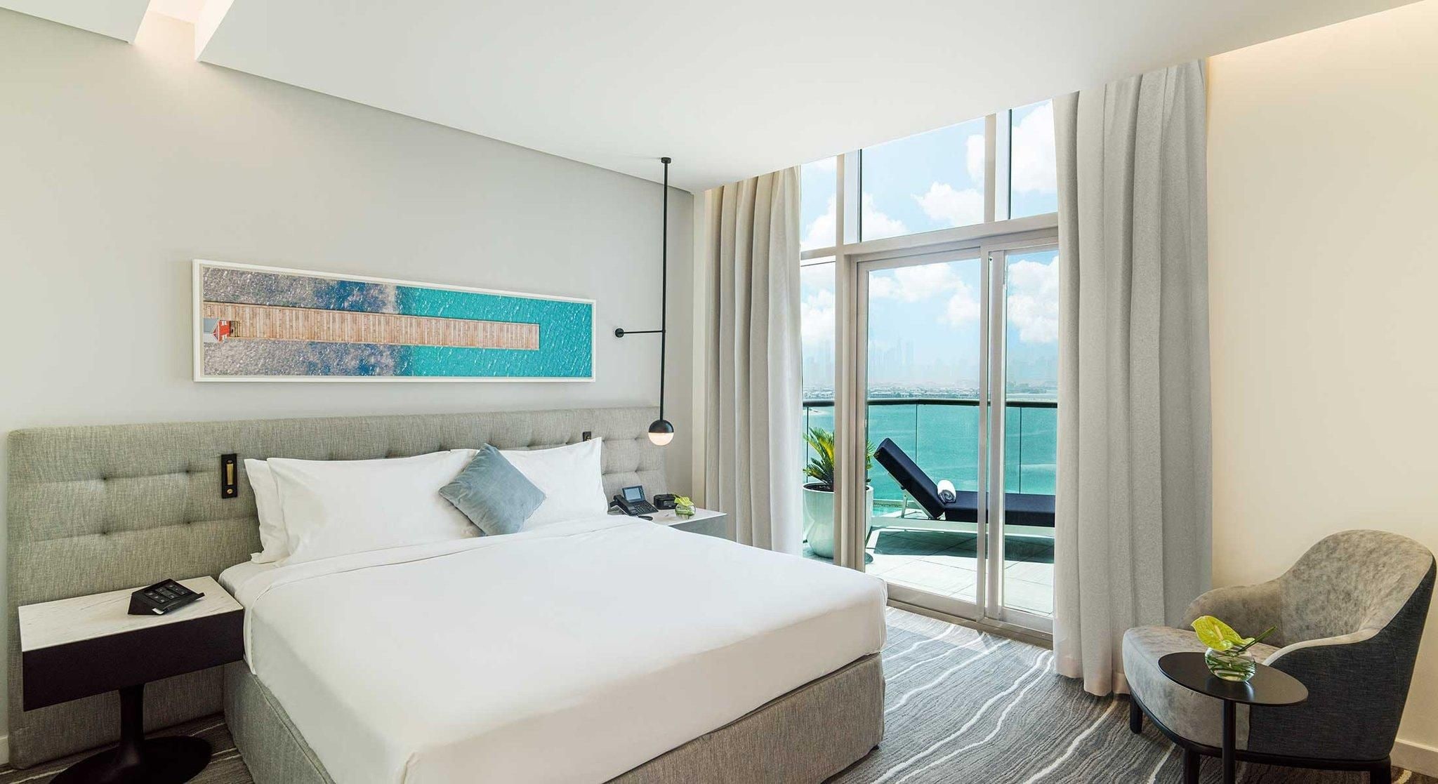 Une chambre d’hôtel comprenant un lit, une table de chevet, une lampe, une chaise et une grande fenêtre donnant sur la mer.